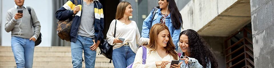 Studierende gehen Treppen herunter, unterhalten sich und zwei Frauen zeigen sich etwas auf Ihrem Handy